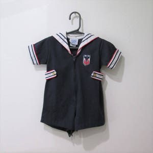 May include: Navy blue romper with a sailor collar, white and red striped sleeve cuffs, and decorative pocket flaps. The romper features a zipper closure and a small embroidered emblem on the chest. The garment is hanging on a black hanger.