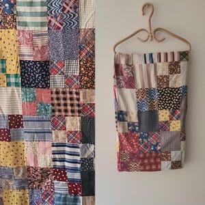 May include: A patchwork quilt featuring a variety of fabric squares in diverse colors and patterns, including stripes, plaids, florals, and polka dots. The quilt is displayed hanging from a light-colored wooden hanger against a plain wall.