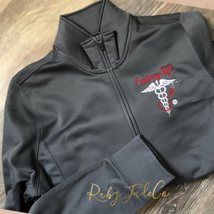 May include: A gray zippered jacket with a red and white embroidered medical symbol and the text "Courtney RN" on the front.