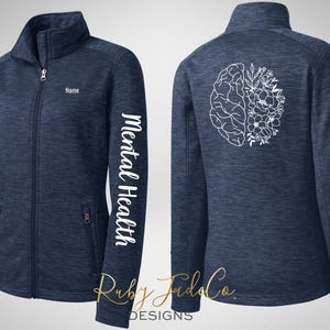 May include: Navy blue zip-up jacket with the words "Mental Health" printed vertically on the left sleeve. The back of the jacket features a white graphic of a brain with floral elements. The jacket has a high collar and two front pockets.