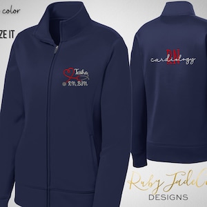 May include: Navy blue zip-up jacket with a red heart and stethoscope design on the left chest. The text "Tasha RN, BSN" is embroidered below the design. The back of the jacket has the text "cardiology" embroidered in white.