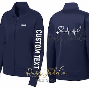 May include: Navy blue fleece zip-up jacket with a white stethoscope and heartbeat graphic on the back. The left sleeve has the text "CUSTOM TEXT" in white. The jacket has a zipper closure and two pockets.