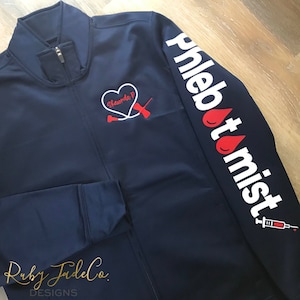 May include: A navy blue zippered jacket with a red heart and a stethoscope embroidered on the left chest. The right sleeve has the word "Phlebotomist" in white with a red blood drop and a syringe.