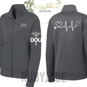 May include: A dark gray zip-up jacket with a white heart beat and stethoscope graphic on the back. The left sleeve has a white caduceus symbol and the text "DOU" and the right sleeve has the text "Nicole BSN, RN".