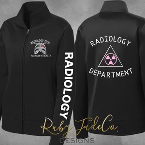 May include: Black zippered jacket with white text that reads "RADIOLOGY TECH" on the left chest and "RADIOLOGY DEPARTMENT" on the back. The left sleeve has the word "RADIOLOGY" printed vertically in white. A white outline of a ribcage with a pink heart is on the left chest.