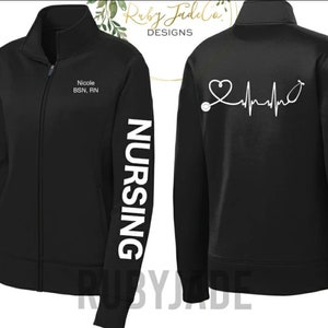 May include: Black zippered jacket with white text on the front and sleeve. The front text reads "Nicole BSN, RN". The sleeve text reads "NURSING". The back of the jacket has a white graphic of a stethoscope with a heart and a heartbeat line.