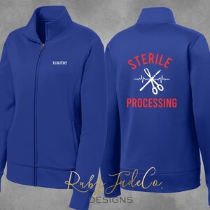 May include: Royal blue fleece jacket with a white zipper and a white embroidered name tag on the left chest. The back of the jacket has a white graphic of crossed surgical scissors with a red heartbeat and the text "STERILE PROCESSING" in red.