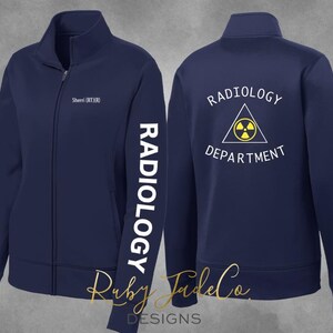 May include: Navy blue zip-up jacket with white text that reads "RADIOLOGY" down the left sleeve. The back of the jacket has the words "RADIOLOGY DEPARTMENT" above a yellow and black radiation symbol. The jacket also has the name "Sherri (RT)(R)" on the front.