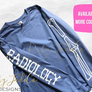 May include: A blue long-sleeve t-shirt with a white skeleton arm design on the sleeve and the word "RADIOLOGY" spelled out in white bones. The text "AVAILABLE MORE COLORS" is in a pink box in the upper right corner of the image.