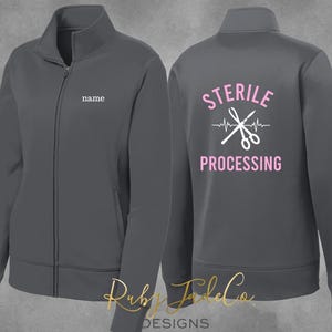 May include: A dark gray zippered jacket with a white embroidered design on the back that says "STERILE PROCESSING" with a heartbeat line and a pair of scissors. The left side of the jacket has a blank space for a name.