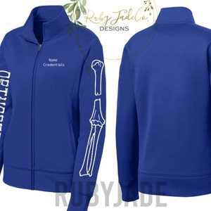 May include: A royal blue zip-up jacket with the word "ORTHOPEDICS" in white block letters down the left sleeve. The right sleeve features a white skeletal arm graphic. The jacket has a mock neck and a zipper closure. The text "Name Credentials" is above the zipper.