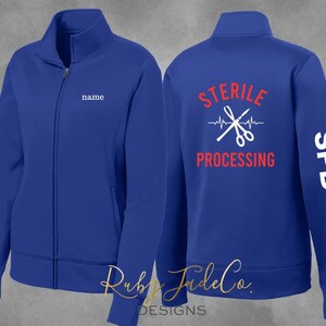 May include: A royal blue zippered jacket with a white embroidered name tag on the left chest. The back of the jacket has a white graphic of scissors and a heartbeat with the text "STERILE PROCESSING" in red. The right sleeve has the text "SPD" in white.