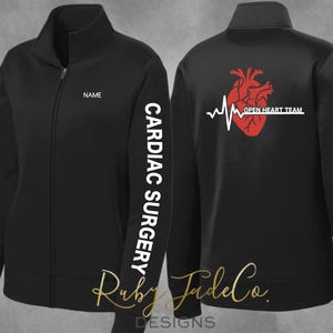 May include: Black zip-up jacket with "CARDIAC SURGERY" printed vertically on the left sleeve. The back features a red heart graphic with a white EKG line and the text "OPEN HEART TEAM". The jacket has a high collar and a full-length zipper.