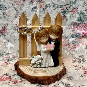 May include: A wooden figurine of a bride and groom standing in front of a white picket fence. The bride is wearing a white dress and a veil, and the groom is wearing a black suit. They are holding a pink heart between them. The figurine is on a wooden base.