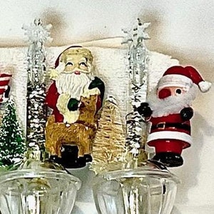 May include: Two white night lights with clear glass shades and a switch on the side. Each night light features a Christmas-themed figurine. One figurine is a Santa Claus holding a reindeer, and the other is a Santa Claus with a white beard and red hat.