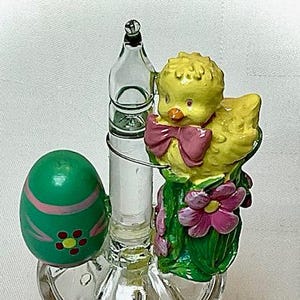 May include: An Easter-themed night light featuring a yellow chick with a pink bow, a green egg with pink stripes, and a clear glass bulb. The base is white with a red berry design. The night light is designed with a clear glass bulb and a metal wire.