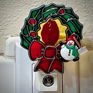 May include: A Christmas-themed nightlight plug-in with a green and red wreath, a red bow, and a snowman holding a small shovel. The wreath has a yellow center with a red and white candy cane.