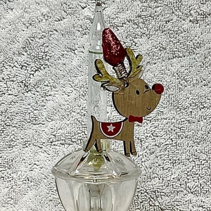 May include: A clear glass night light with a reindeer figurine on top. The reindeer is brown with a red nose and a red bow tie. The night light has a white base with an on/off switch.