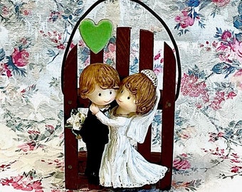 May include: A wedding cake topper featuring a bride and groom figurine standing in a wooden archway with a green heart above them. The archway is decorated with a black wire heart.