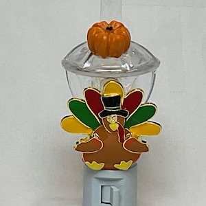 May include: Thanksgiving-themed night light featuring a colorful turkey with a black top hat and a small orange pumpkin on top. The turkey has red, green, and yellow feathers. The night light is made of plastic.