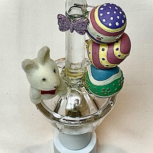 May include: A decorative Easter-themed night light. The night light features a clear glass base with a small white bunny wearing a red bow tie. Above the base is a clear glass column with a purple glitter butterfly and three colorful Easter egg ornaments.
