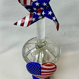 May include: A clear glass night light with a red, white, and blue American flag pinwheel on top and a heart-shaped American flag on the front. The night light has a white electrical cord and plug.