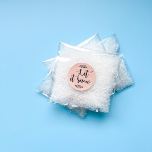 May include: Three clear plastic bags filled with white granules, possibly bath salts or decorative snow, are stacked on a light blue surface. A round, pink sticker with the words "Let it snow" in elegant script is affixed to one bag.