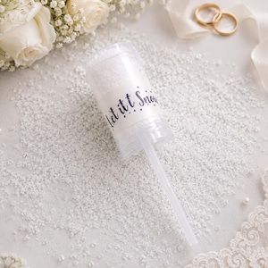 May include: A clear plastic push-pop filled with white confetti, with the words "Let it Snow" in a dark blue script. The pop-up is on a white surface scattered with confetti, with white roses and wedding rings in the background.