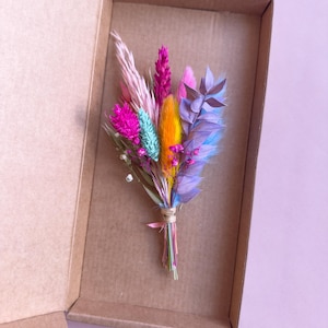 May include: A small bouquet of dried flowers in a cardboard box. The flowers are pink, purple, blue, yellow, and white.