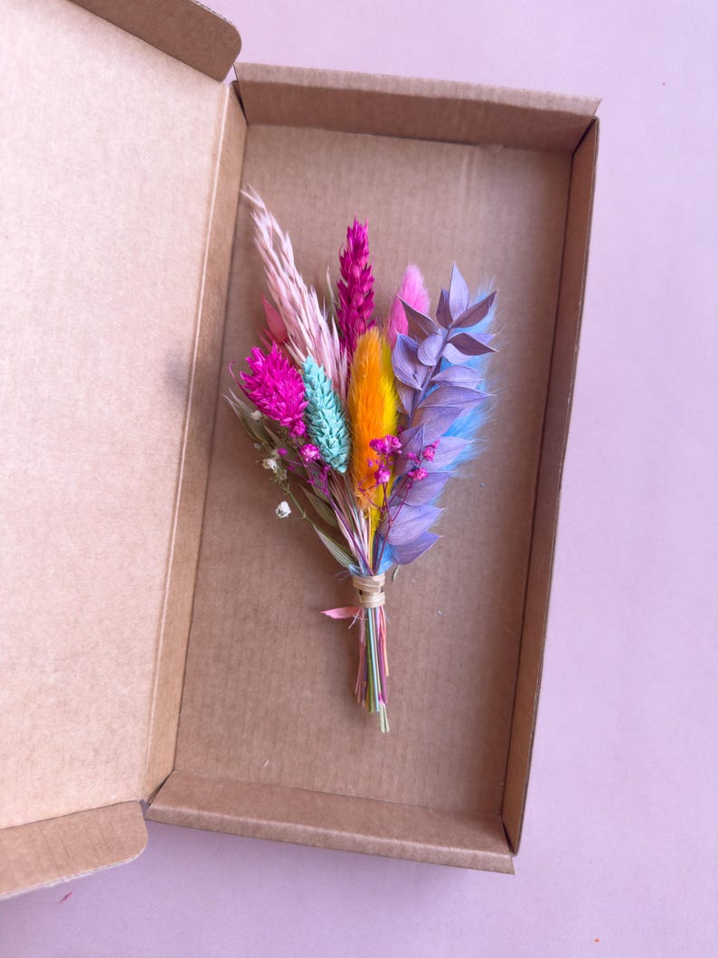 May include: A small bouquet of dried flowers in a cardboard box. The flowers are pink, purple, blue, yellow, and white.