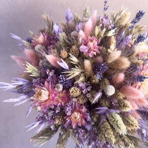 May include: A dried flower bouquet featuring a mix of purple, pink, and beige flowers and grasses. The bouquet is arranged in a circular shape and showcases a variety of textures.