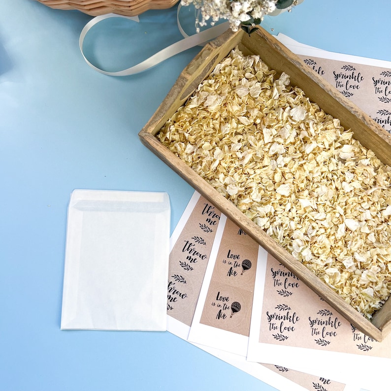 Op de afbeelding: Een houten doos gevuld met witte bloemblaadjes, samen met verschillende bruine papieren kaartjes met de tekst "Throw me", "Love is in the Air" en "Sprinkle the Love".
