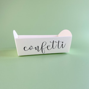 May include: White cardboard box with a handle on each side. The box is labeled "confetti" in black cursive writing.