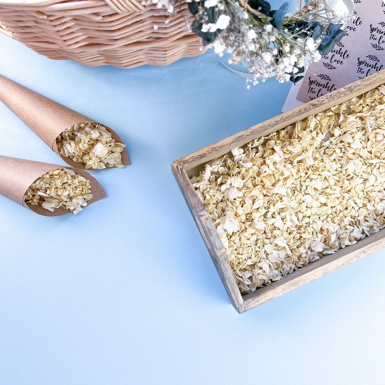 Op de afbeelding: Een houten doos gevuld met witte bloemblaadjes, met twee bruine papieren kegels gevuld met dezelfde bloemblaadjes. De kegels staan op een lichtblauw oppervlak. De tekst op het bruine papier zegt "Sprinkle the love".