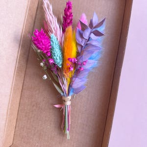May include: A small bouquet of dried flowers in a cardboard box. The flowers are pink, purple, blue, orange, and green.