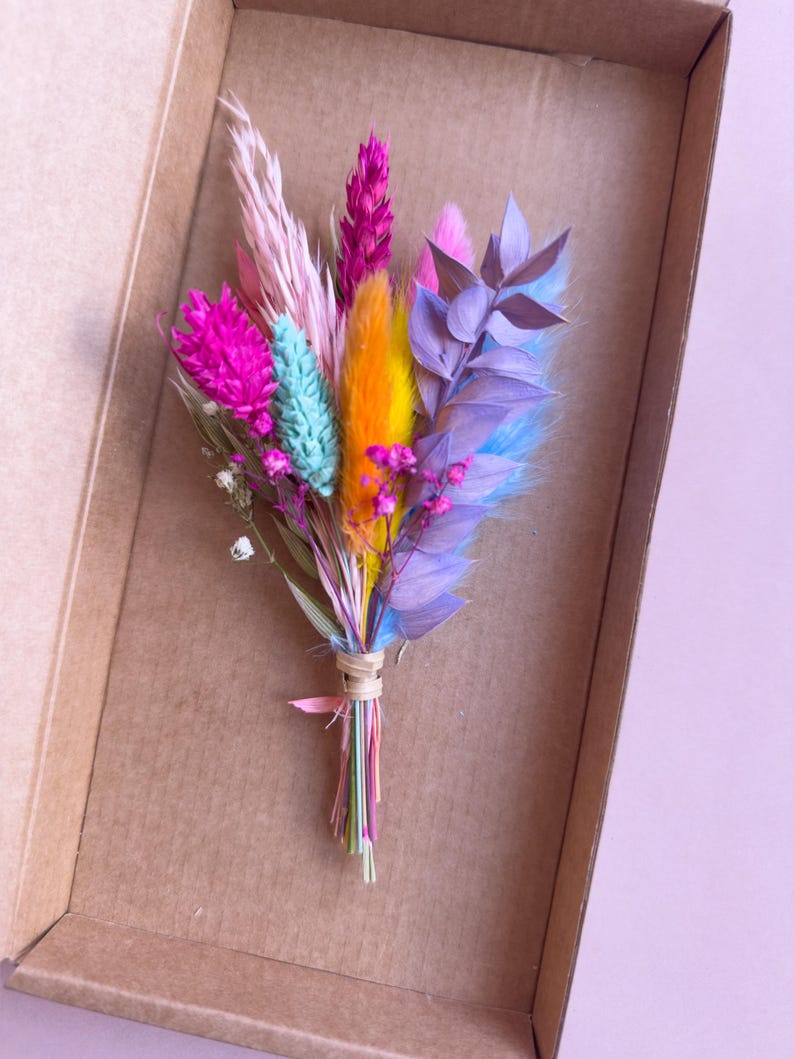 May include: A small bouquet of dried flowers in a cardboard box. The flowers are pink, purple, blue, orange, and green.