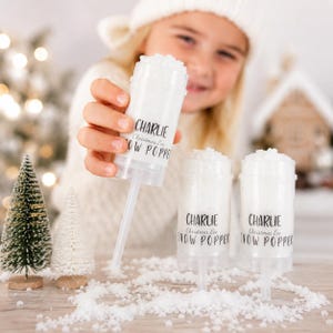 Op de afbeelding: Drie doorzichtige plastic sneeuwpoppers gevuld met witte confetti, met de tekst "CHARLIE Christmas Eve SNOW POPPER" erop gedrukt. Twee kleine kerstbomen en verspreide confetti zijn ook zichtbaar.