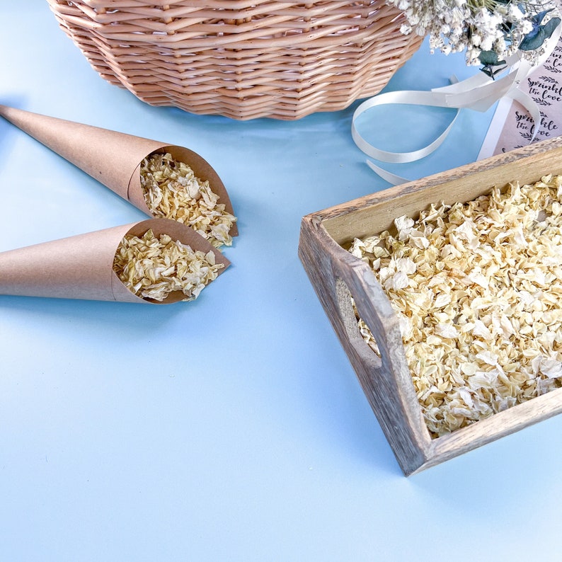 Op de afbeelding: Twee bruine papieren kegels gevuld met gedroogde bloemblaadjes, een houten dienblad met gedroogde bloemblaadjes en een wit lint op een lichtblauwe achtergrond. Het lint heeft een label met de tekst "Sprinkle the Love".