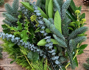 Op de afbeelding: Een feestelijke krans bestaande uit blauwgroene dennentakken, glanzende groene hulstbladeren, zilverkleurige eucalyptusstengels en trossen kleine geelgroene bessen. Het loof is gelaagd om een dichte, ronde botanische compositie te vormen.