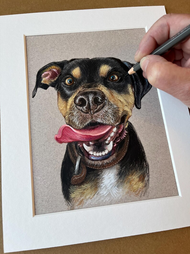 Puede incluir: Dibujo a l&aacute;piz de color de un perro con expresi&oacute;n feliz. El perro tiene pelaje negro, marr&oacute;n y beige, una lengua rosa y un collar marr&oacute;n. La obra de arte est&aacute; en un passe-partout blanco.