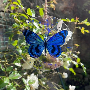 May include: A blue butterfly painted on a clear round glass ornament with a string for hanging. The ornament has blue and black paint splatters on it.