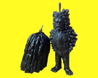 Op de afbeelding: Twee zwarte wax kaarsen, een in de vorm van een monster en de andere in de vorm van een boom. De monsterkaars heeft een getextureerd oppervlak en een woeste uitdrukking.