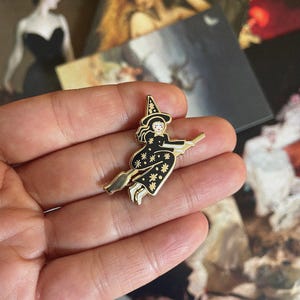 Könnte beinhalten: Ein goldfarbener Emaille-Pin mit einer Hexe auf einem Besen. Die Hexe trägt ein schwarzes Kleid mit weißen Sternen und einen spitzen Hut. Der Pin wird in einer Hand gehalten, im Hintergrund sind Vintage-Kunstdrucke zu sehen.