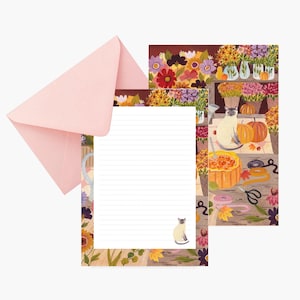 Peut inclure: Un ensemble de papeterie comprenant une enveloppe rose clair et deux feuilles de papier ligné décorées d'illustrations d'un chat siamois, de citrouilles, de feuilles d'automne et de fleurs colorées en vases, disposées sur une surface en bois.