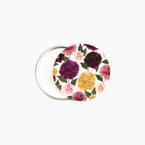 May include: A round compact mirror with a floral pattern. The design showcases dahlias in burgundy, pink, yellow, and white, with green leaves. The mirror is open, revealing a reflective surface.