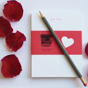 Può includere: Un blocco note con una striscia rossa, un cuore bianco e un'illustrazione di macchina da scrivere, con il testo "love note" e "Calla Design Vancouver". Una matita grigia è appoggiata sul blocco note, circondata da petali di rose rosse.