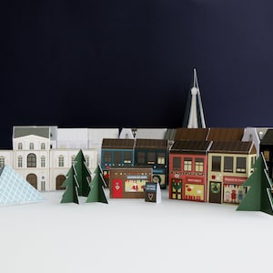 Puede incluir: Una escena de pueblo en miniatura de papel con edificios, árboles y una pirámide. Los edificios son de varios colores y estilos, algunos con escaparates. Los árboles de papel verde están intercalados en la escena, creando un ambiente festivo.