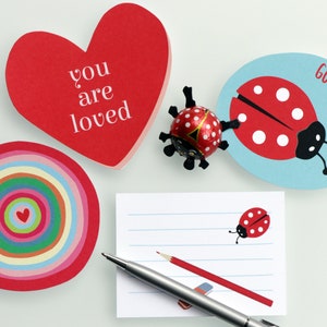 Pode incluir: Uma coleção de artigos de papelaria, incluindo um cartão vermelho em forma de coração com o texto "you are loved", um cartão circular com um design de arco-íris e um cartão oval azul com uma ilustração de joaninha e o texto "good luck". Inclui bloco de notas, lápis e caneta.