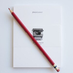 Può includere: Una matita rossa è appoggiata in diagonale su una carta bianca con il testo "#whatever" e un'illustrazione di macchina da scrivere. La carta ha anche il testo "CB DESIGN MAKER". La matita ha una fascia blu e oro e una gomma rosa.