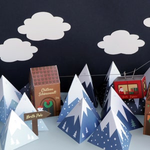 以下が含まれることがあります： 紙製の山、雲、建物を使ったお祝いのクリスマスアドベントカレンダーのシーン。 「サンタエクスプレス」の文字が入った赤いロープウェイが山頂の間に吊り下げられています。 シーンには「北極」と書かれた看板が含まれています。